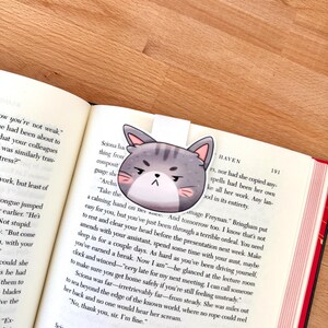 May include: A magnetic bookmark featuring a cartoon cat head with grey stripes, pink inner ears, and a grumpy facial expression. The bookmark is clipped onto the top edge of an open book page with printed text, showing a portion of a novel.