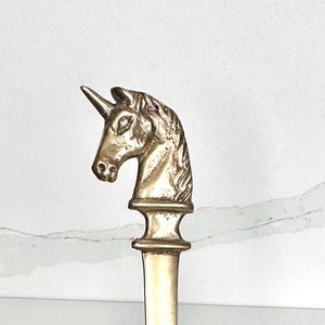 May include: A brass letter opener with a unicorn head design. The unicorn head is detailed with a horn, mane, and facial features. The letter opener is held in a hand against a white background.