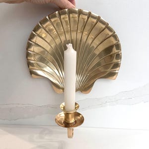 May include: A gold-coloured seashell-shaped wall sconce with a white candle. The sconce has a detailed, fan-like design and a circular candle holder. The candle is white and unlit, adding a touch of elegance to the decor.