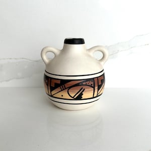 Puede incluir: Un pequeño jarrón de cerámica color crema con dos asas y un borde negro. El jarrón presenta un diseño geométrico en tonos marrones y negros en el centro. La parte superior del jarrón es negra.