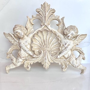 May include: A decorative wall plaque featuring two winged cherubs flanking a central scalloped shell motif. The piece is finished in an off-white, distressed paint with visible brown undertones, showcasing intricate carved details and scrollwork.