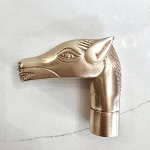 Op de afbeelding: Een gepolijste koperen handgreep in de vorm van een paardenkop, met gedetailleerde gravures voor het oog, het oor en de manen. De paardenkop is naar links gericht, met een cilindrische basis aan de onderkant voor bevestiging aan een wandelstok.