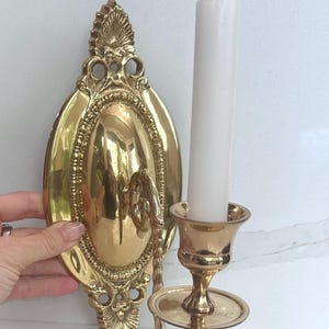 Pode incluir: Um candeeiro de parede em latão polido com uma placa traseira oval com detalhes em relevo decorativos e um braço curvo que suporta um suporte para vela. Uma vela cónica branca, alta e fina, está inserida no suporte.