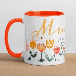 May include: White ceramic mug with an orange rim and handle. The mug features a floral design with colorful flowers and bees. The word "Mum" is written in a yellow script font.