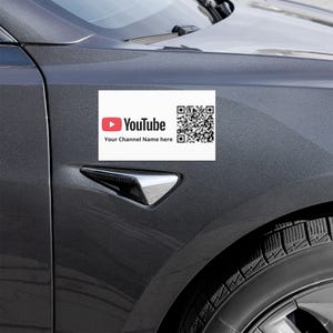 May include: A white rectangular car magnet with the YouTube logo and a QR code. The YouTube logo features a red play button and the word "Youtube" in white. Below the logo is the text "Your Channel Name here". The magnet is affixed to a gray car.