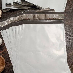 May include: A stack of white poly mailers with a silver and black seal. The mailers are flat and have a smooth surface.