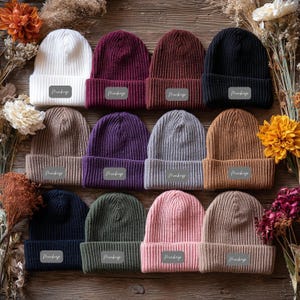 Op de afbeelding: Een verzameling geribbelde gebreide mutsen in diverse kleuren, waaronder wit, bordeauxrood, bruin, zwart, beige, paars, grijs en roze. Elke muts heeft een omgeslagen rand en een klein rechthoekig label met de tekst "Mockup". De mutsen zijn geplaatst op een houten ondergrond.