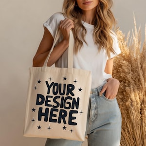 May include: A cream-coloured tote bag featuring the text "YOUR DESIGN HERE" in navy blue, with star motifs. The bag is held by a person wearing a white t-shirt and blue jeans.
