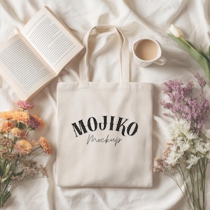 May include: A cream-coloured tote bag with the text "MOJIKO Mockup" in black, set on a white sheet. An open book, a cup of coffee, and flowers in shades of orange, purple, and white are arranged around the bag, creating a cosy, aesthetic scene.