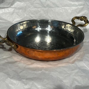May include: A copper-colored pan with a hammered interior and brass handles. The pan has a rounded shape and two ornate handles on either side. The interior is a shiny silver color.