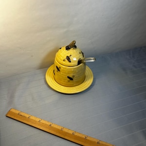 May include: A yellow ceramic honey pot with a lid and a small spoon, decorated with black bee motifs. The pot sits on a matching saucer. The honey pot is approximately 4 inches tall.