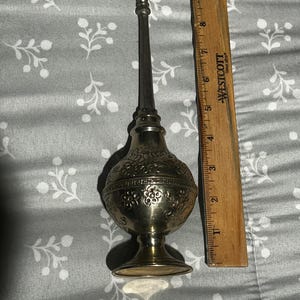 May include: A silver-colored, ornate perfume bottle with a long, slender applicator. The bottle has a bulbous body with floral and scrollwork designs. A wooden ruler is next to the bottle, measuring approximately 6 inches in height.