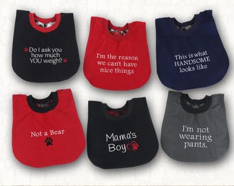 May include: Six baby bibs in black, red, navy blue, and gray with white embroidered text. Phrases include Do I ask you how much YOU weigh?, I'm the reason we can't have nice things, This is what HANDSOME looks like, Not a Bear, Mama's Boy, and I'm not wearing pants.