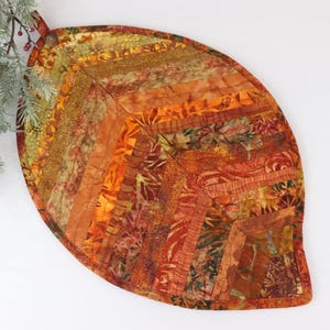May include: A quilted leaf-shaped table runner in shades of orange, brown, and green. The runner is made of fabric scraps sewn together in a patchwork design.
