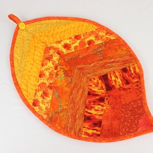 May include: A quilted leaf-shaped hot pad in shades of orange and yellow. The pad is made of patchwork fabric and has a button on the top.