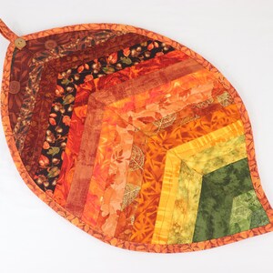 May include: A quilted leaf-shaped hot pad in shades of orange, red, yellow, and green. The pad is made of fabric with a variety of patterns, including floral and geometric designs.