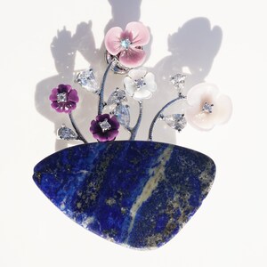 Può includere: Un ciondolo d'argento con un gruppo di fiori colorati su una base di pietra lapislazzuli. I fiori sono realizzati in smalto rosa, bianco e viola con strass trasparenti.