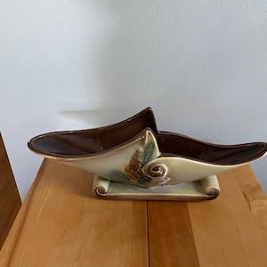 May include: A ceramic boat-shaped planter with a glossy dark brown interior and a cream-colored exterior. The exterior features a raised, textured relief of a pinecone and needles in shades of brown and green, set on a rectangular base.