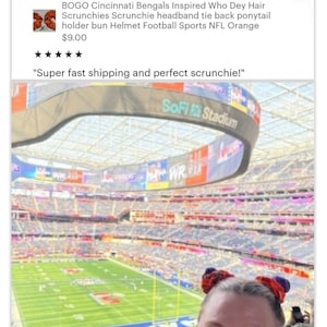 May include: A woman wearing a bright orange shirt with two orange and purple scrunchies in her hair, smiling at the camera. The woman is sitting in a stadium with a large screen displaying "SoFi Stadium".