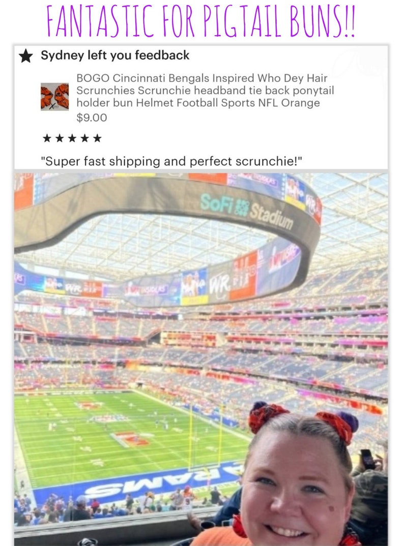 May include: A woman wearing a bright orange shirt with two orange and purple scrunchies in her hair, smiling at the camera. The woman is sitting in a stadium with a large screen displaying "SoFi Stadium".