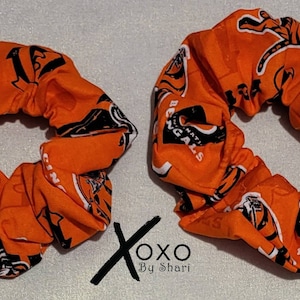 May include: Two orange and black scrunchies with the Cincinnati Bengals logo and tiger stripes. The scrunchies are made of fabric and have a gathered look.