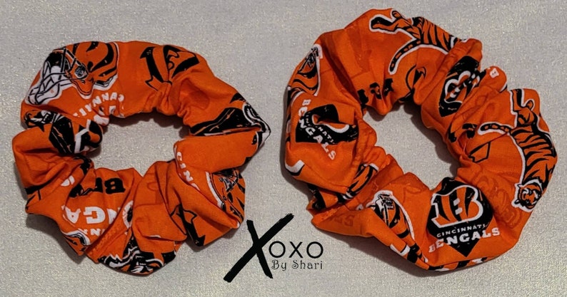 May include: Two orange and black scrunchies with the Cincinnati Bengals logo and tiger stripes. The scrunchies are made of fabric and have a gathered look.