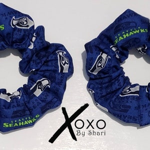 May include: Two blue scrunchies with a white and green Seattle Seahawks logo pattern. The scrunchies are made of fabric and have a gathered look.