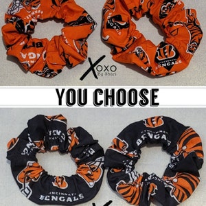 May include: Two orange and black scrunchies with the Cincinnati Bengals logo and tiger stripes. The scrunchies are made of fabric and have a stretchy elastic band.