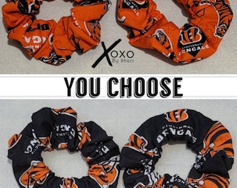 May include: Two orange and black scrunchies with the Cincinnati Bengals logo and tiger stripes. The scrunchies are made of fabric and have a stretchy elastic band.