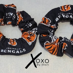 May include: Two black and orange scrunchies with the Cincinnati Bengals logo. The scrunchies are made of fabric and have a white background with the Bengals logo in orange and black.