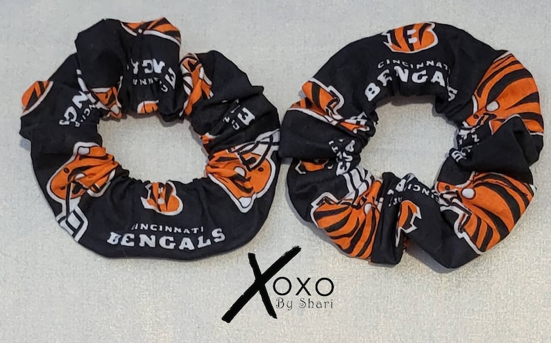 May include: Two black and orange scrunchies with the Cincinnati Bengals logo. The scrunchies are made of fabric and have a white background with the Bengals logo in orange and black.