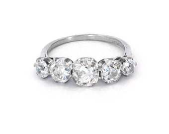 May include: A silver ring featuring five clear, round-cut gemstones. The gemstones are set in a row across the top of the ring, with the center stone being the largest. The ring is set against a white background.
