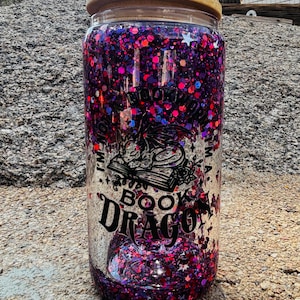 May include: A clear glass tumbler with a bamboo lid, filled with purple and pink glitter. The exterior features a black graphic of a dragon resting on an open book, with the text "I'M NOT A BOOKWORM I'M A BOOK DRAGON" printed in black lettering.