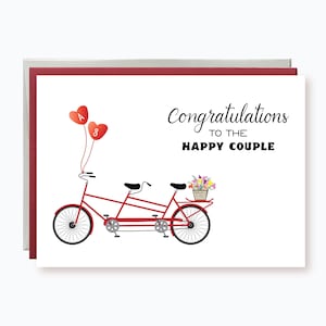 May include: A white card with the text "Congratulations to the Happy Couple" and a red tandem bicycle with a basket of flowers on the back. Two red hearts with the letters "A" and "3" are floating above the bicycle.