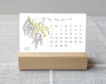 May include: A rectangular desk calendar for June 2027 featuring a watercolor illustration of purple wisteria flowers with green leaves on the left side. The calendar is printed on white cardstock and held upright by a rectangular wooden block stand.