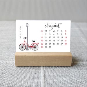 May include: A rectangular desk calendar for August 2027 printed on white cardstock, featuring a red bicycle illustration next to a gray street lamp. The calendar is held upright by a rectangular light-colored wooden block stand.
