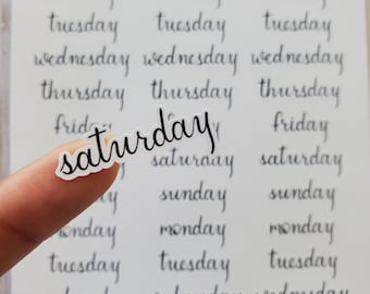 May include: A sheet of clear stickers with the days of the week written in cursive script. The word "saturday" is highlighted with a finger.