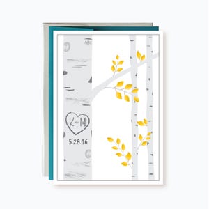 May include: A rectangular greeting card featuring a white background with illustrations of grey birch tree trunks and bright yellow leaves. A heart shape is carved into the main trunk containing the letters K+M and the date 5.28.16 below it.