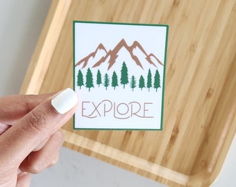 May include: A white sticker with a green border features a brown mountain range with green trees in front of it. The text "EXPLORE" is written in brown letters below the mountains.
