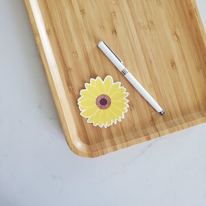 May include: A yellow sunflower sticker on a wooden surface with a white pen.