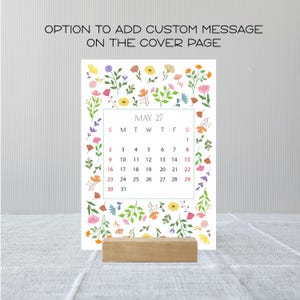May include: A vertical calendar page for May 27 featuring a colorful floral border with various small flowers and green leaves. The text reads OPTION TO ADD CUSTOM MESSAGE ON THE COVER PAGE. The calendar is held upright by a rectangular wooden block.