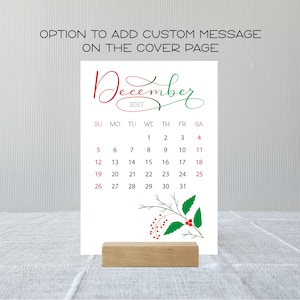 May include: A December 2027 calendar page featuring a red and green script header, a grid of dates, and a botanical illustration of holly leaves and red berries at the bottom. The text reads OPTION TO ADD CUSTOM MESSAGE ON THE COVER PAGE.