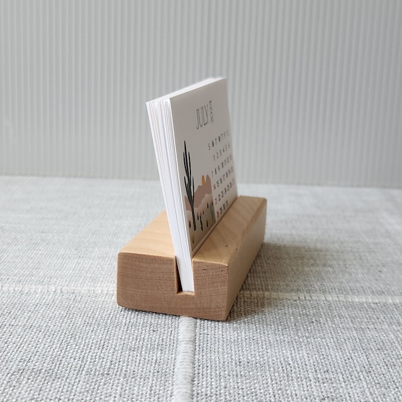 May include: A wooden desk calendar holder with a white card calendar that says "JULY 2023" and features a desert landscape with a cactus and a sunset.