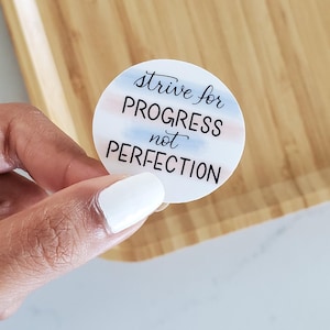 May include: A white sticker with a blue and pink watercolor design. The sticker says "strive for progress not perfection".