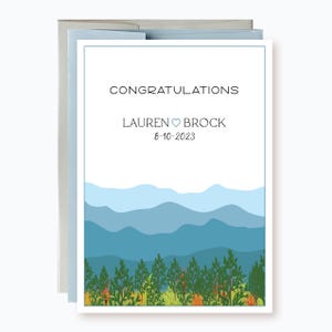 May include: A white card with a blue and green mountain landscape design. The text "Congratulations Lauren ♥ Brock 8-10-2023" is printed on the card.