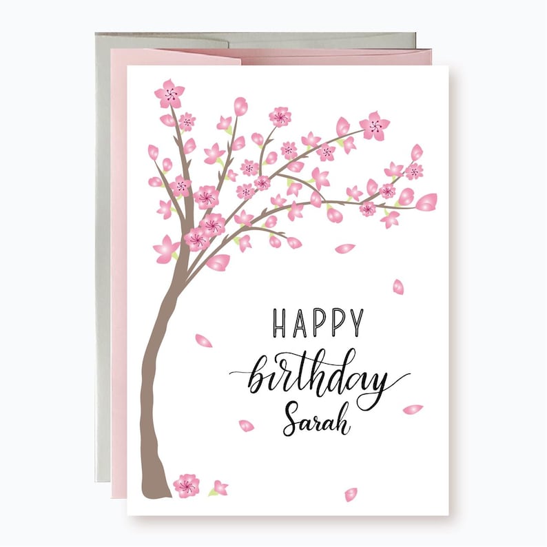 May include: A white card featuring a pink cherry blossom tree design. The text "Happy birthday Sarah" is written in black cursive.