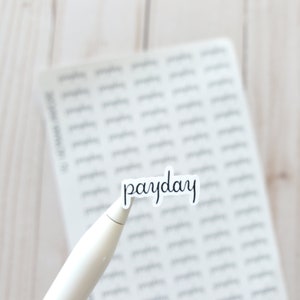 May include: A sheet of stickers with the word "payday" written in cursive on one of the stickers. The stickers are on a white wooden surface.