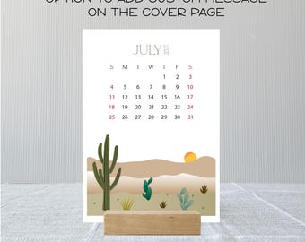 May include: A July 2027 calendar page featuring a desert landscape with cacti and a sun, displayed on a wooden stand. The text at the top reads OPTION TO ADD CUSTOM MESSAGE ON THE COVER PAGE. The calendar grid shows dates in black and red ink.