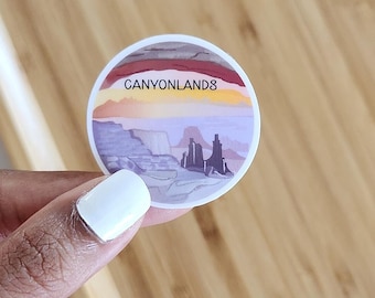 May include: A round sticker featuring a watercolor illustration of Canyonlands National Park. The design includes a sunset sky with the text "CANYONLANDS" above a landscape of mesas and buttes in shades of purple, orange, and gray.
