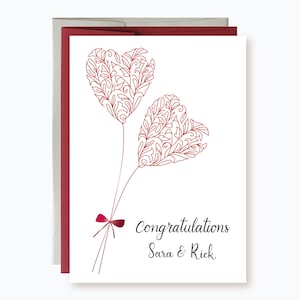 May include: A white card with a red envelope. The card features two red heart-shaped balloons with intricate designs. The text "Congratulations Sara & Rick" is printed in black at the bottom of the card.